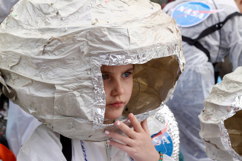 A little girl wears a homemade NASA spacesuit with a large, round helmet made of paper maché.