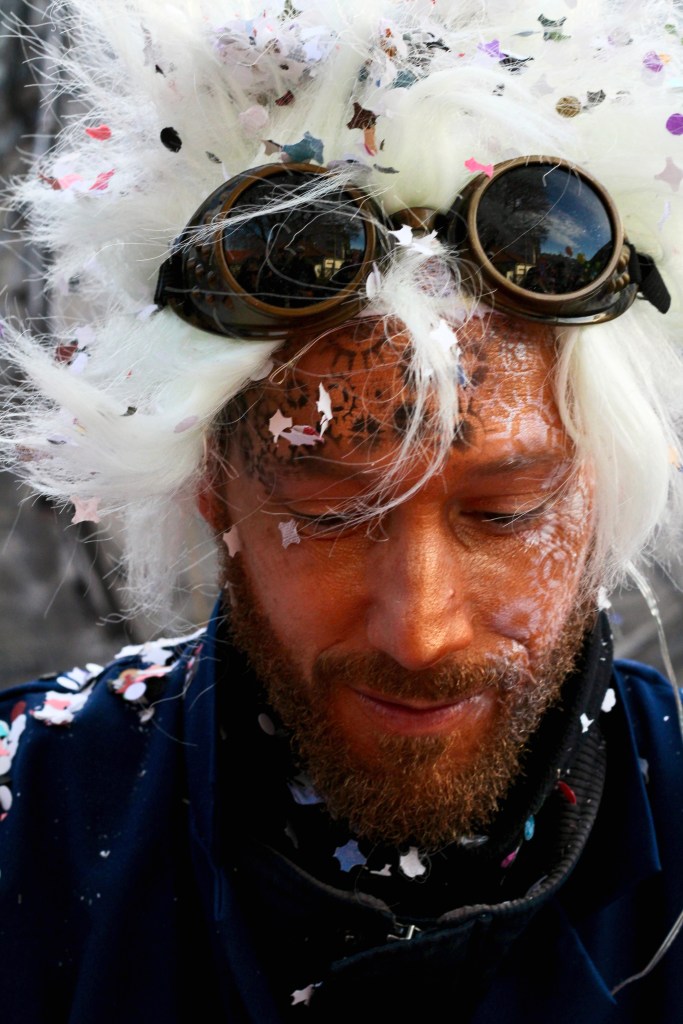 A man with a white wig, goggles on his forehead and face painted gold. 
